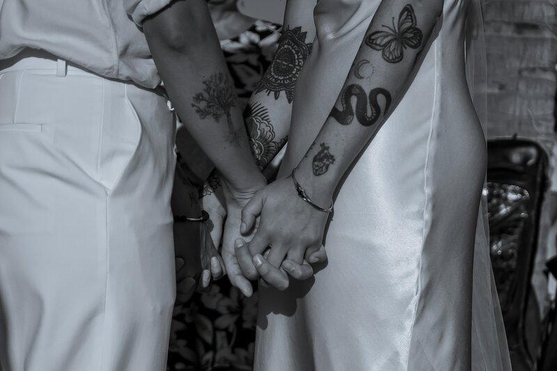 Black and white close-up of two brides holding hands, both with visible tattoos on their arms, one wearing a satin wedding dress.