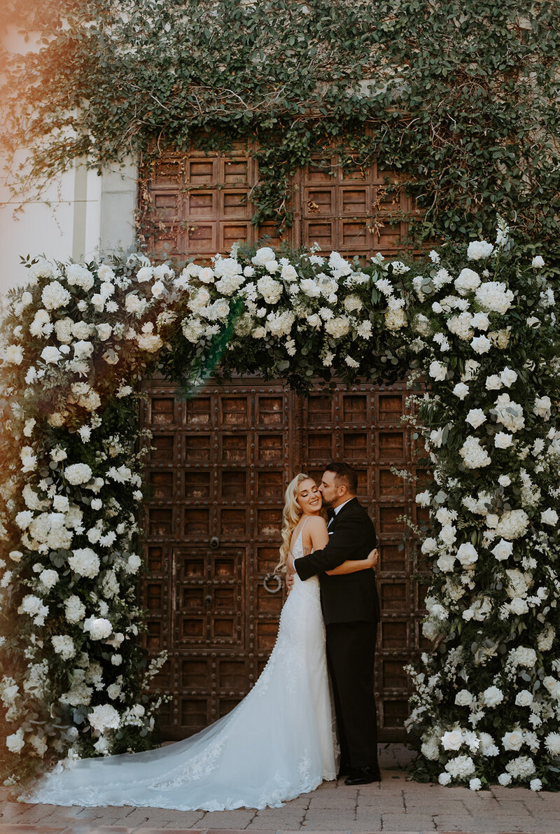 Bride and groom standing beneath large floral arch in front of elegant grand doorway at Arizona wedding