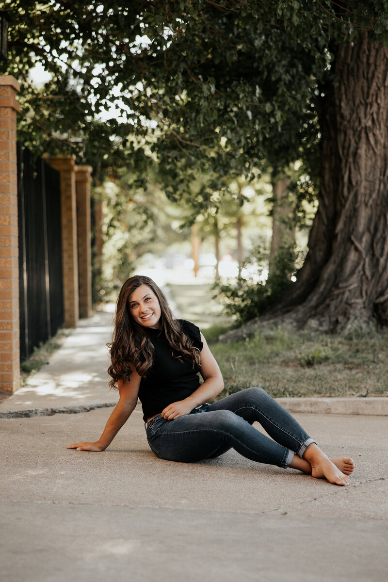 senior sitting on sidewalk wearing a black shirt with denim jeans and she is barefoot.