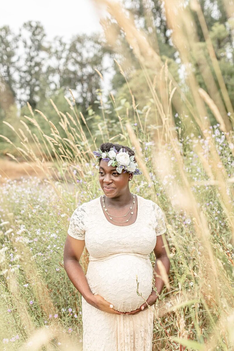 dark skinned pregnant woman holding belly in beautiful lace maternity gown with a floral crown out in nature for maternity portraits in portland oregon