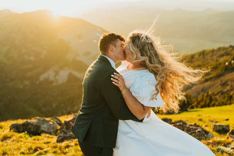 A bridal session in Eastern Idaho where the groom is kissing his bride on top of a mountain during golden hour. The win is blowing the bride's hair and it looks like a snapshot in time.