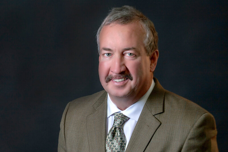 A middle-aged man with short gray hair and a mustache, dressed in a brown suit jacket, white shirt, and patterned tie, is smiling in front of a dark background—an ideal example of professional headshots Jacksonville clients seek.