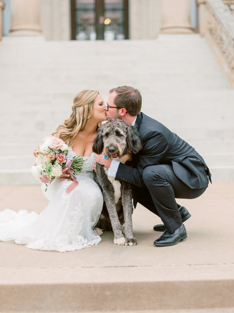 A bride and groom kneeling down to pet their dog and kissing