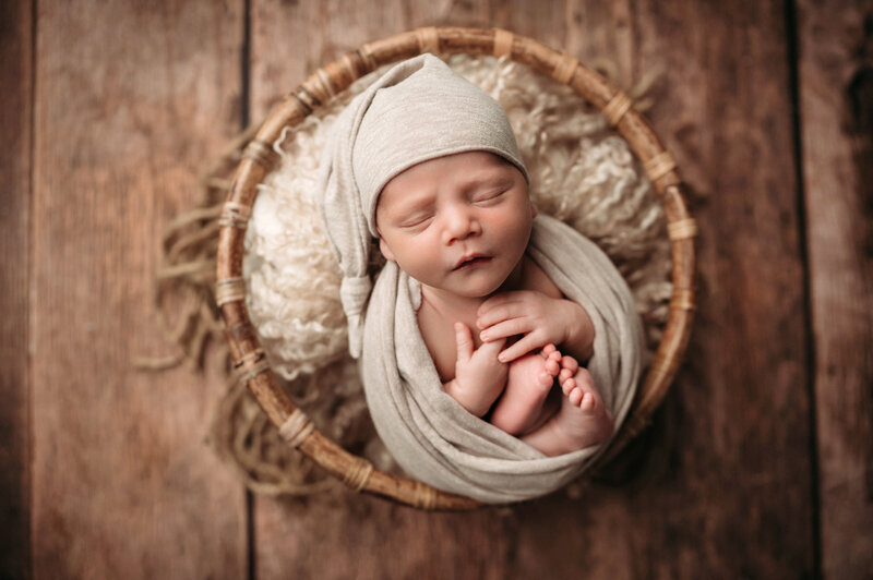 two week old newborn baby boy wrapped in a beige wrap with toes and hands showing and wearing a sleepy cap posed inside round brown bowl over brown flooring