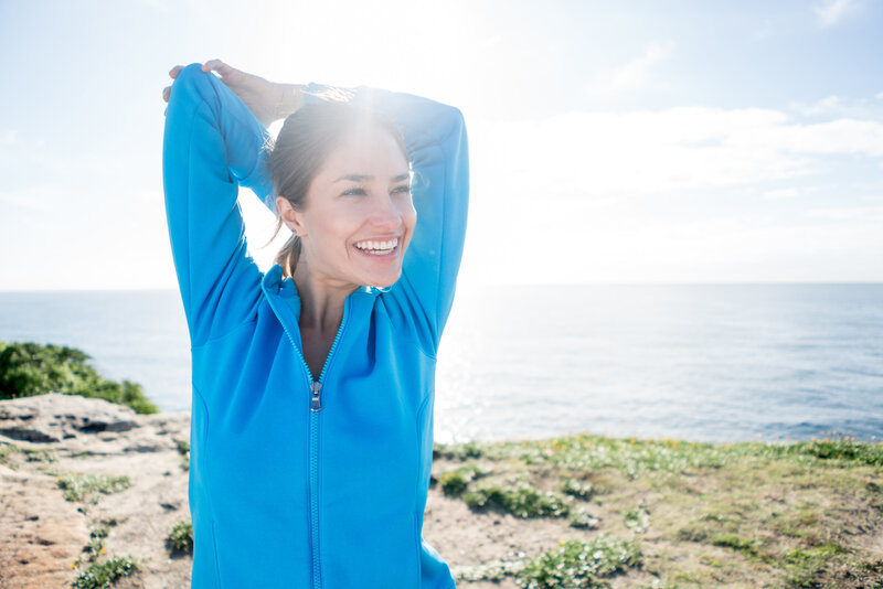 Vibrant young woman stretches arm in blue warm up jacket by the ocean