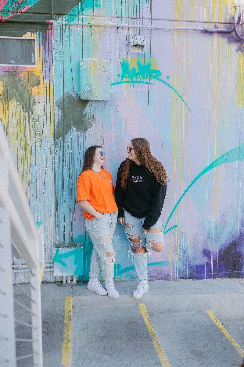 Two friends laughing and standing against a colorful wall.