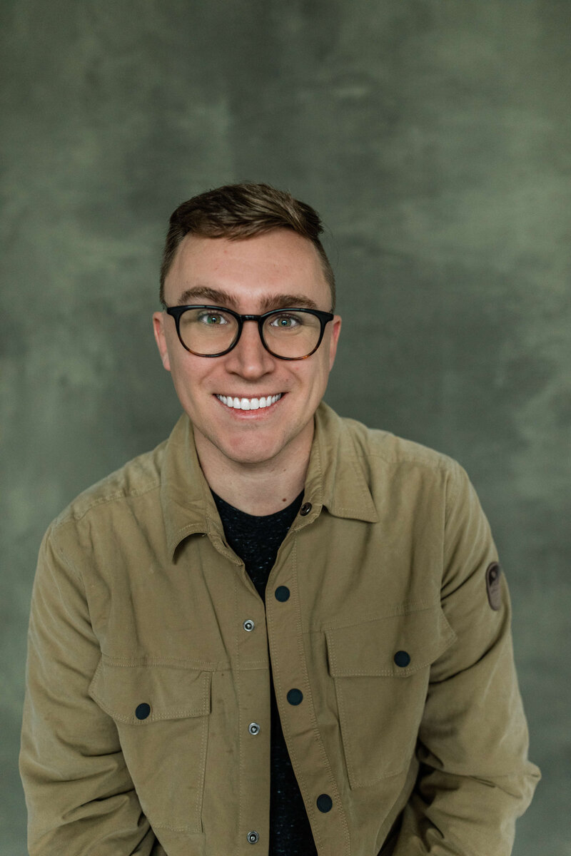 Brunette man smiling for camera wearing black glasses and army green button up shirt
