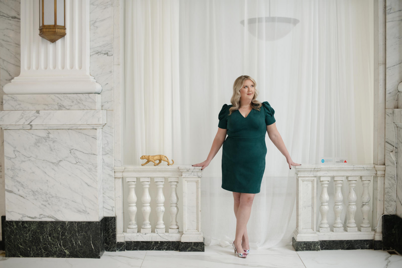 Melissa Lawrence wears teal dress and stands in walkway in front of drapes