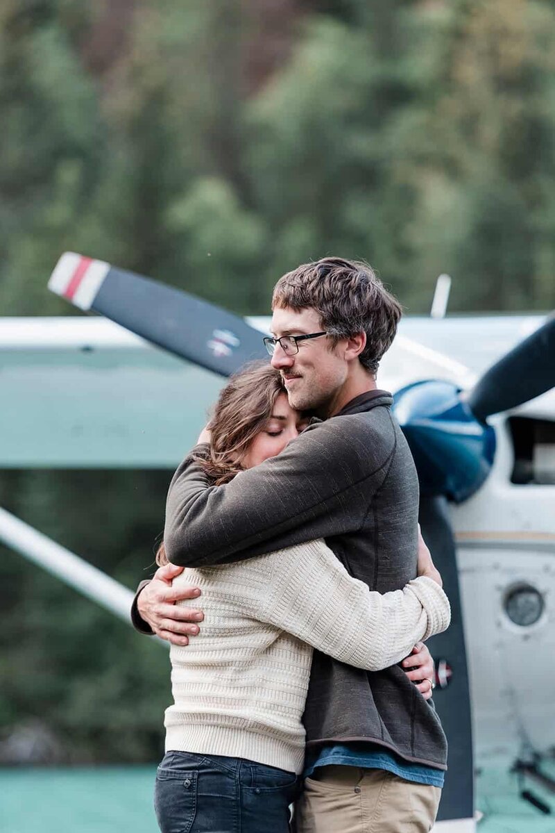 A man and woman embrace tenderly, standing in front of a float plane propeller during their romantic alaska vacation. 