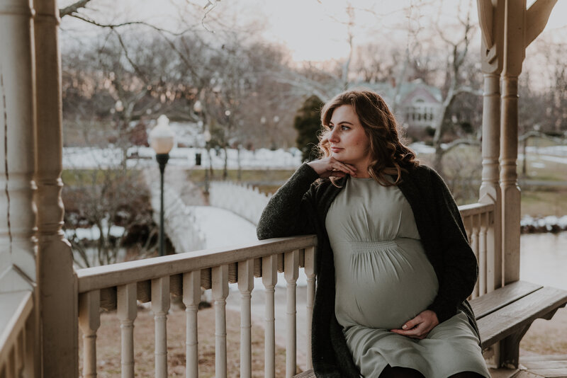 Pregnant mom in black sweater and green dress sitting in gazebo bench looking out to the left while holding belly and having her hand under her chin.