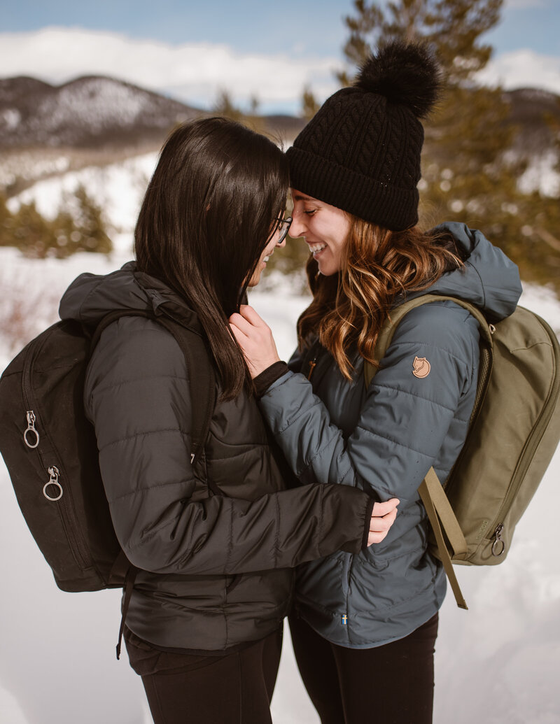 Two women bundled in winter jackets and backpacks stand close together in the snow, smiling and touching foreheads against a mountain backdrop.