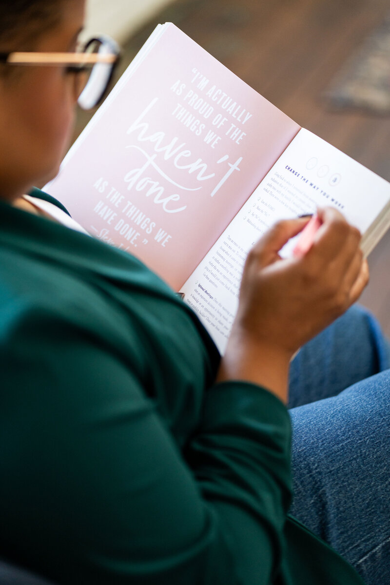 Expert in C-Suite career coaching reading an inspirational book in a casual green outfit, focusing on healing from corporate trauma and helping clients refine their skillsets for 6-figure positions.