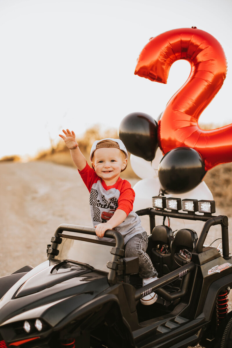 toddler boy celebrates his second birthday in a black jeep with balloons tied to it