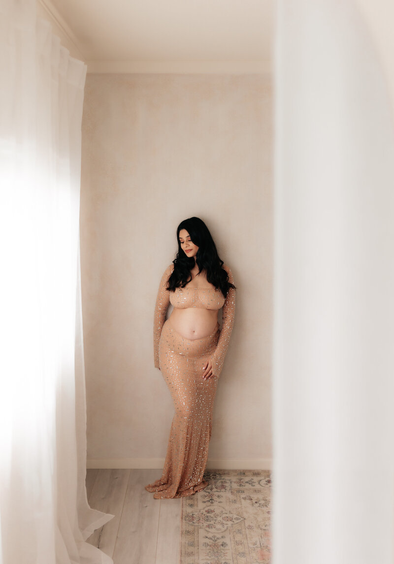 A pregnant mother with beautiful brown hair stands in an all white Newport Beach studio wearing a sheer gown. 