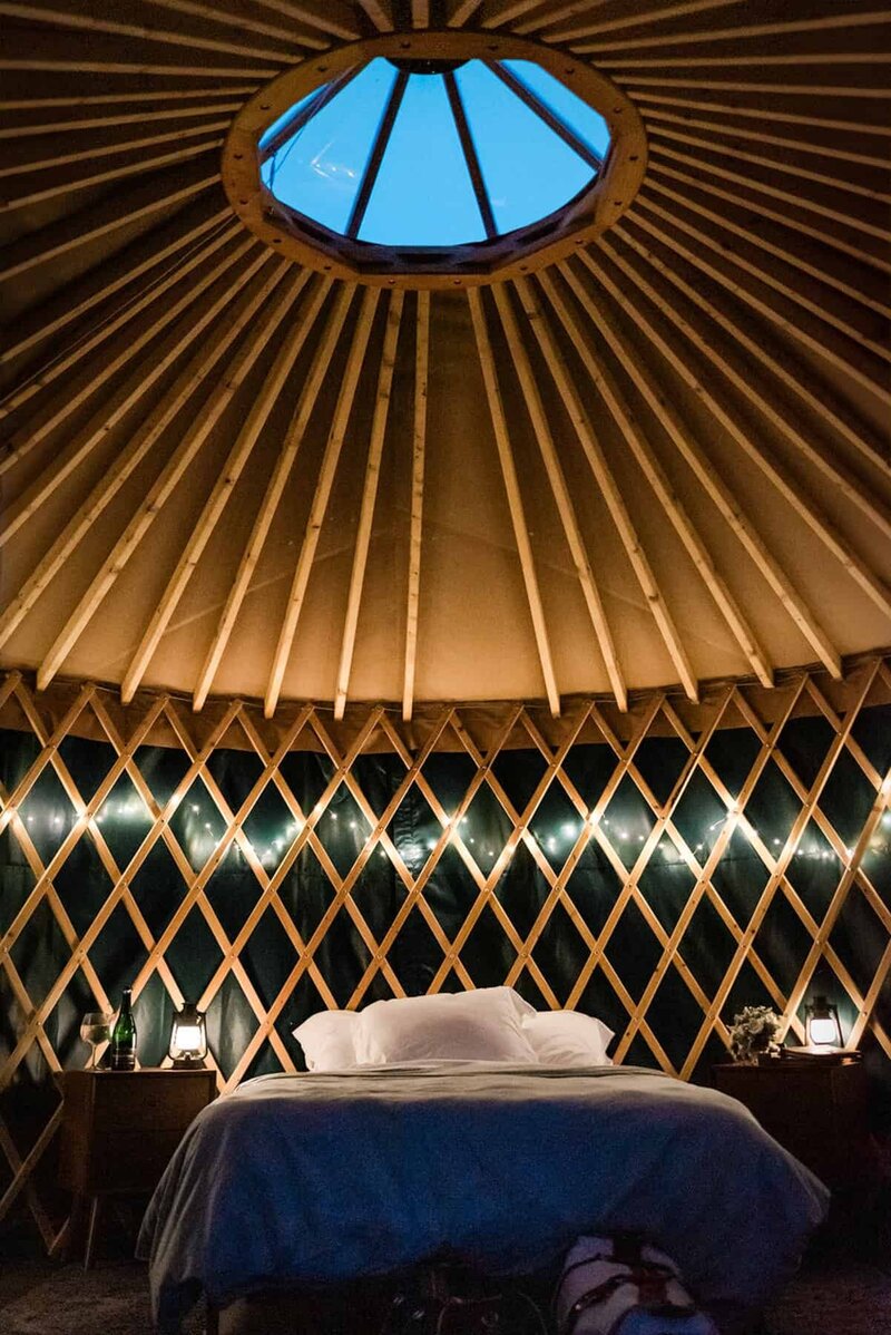 It is dark out, and the glamping yurt with it's wooden interior frame yurt is glowing with warm light from twinkle lights and small lanterns on the decorative tables next to the bed with cozy blankets and pillows that is visible through the open door as the couple inside ends their adventure elopement day.  