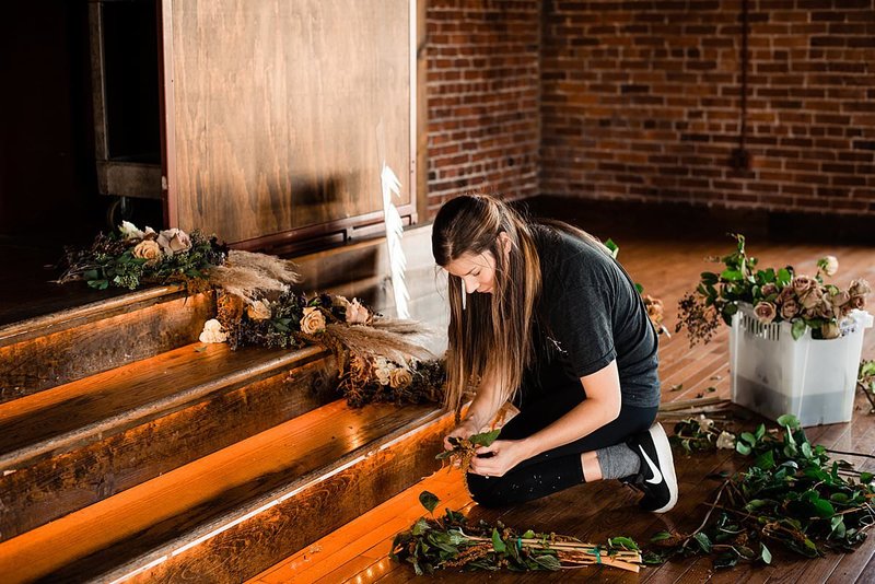 Florist kneeling near wooden stairs cutting stems to arrange for a flower instillation on the staircase