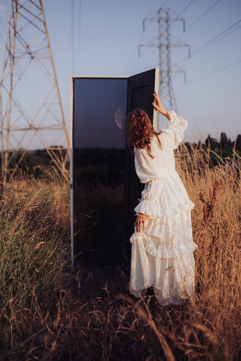 Woman wearing a flowy white dress in a field peeks through a white door with powerlines behind her.