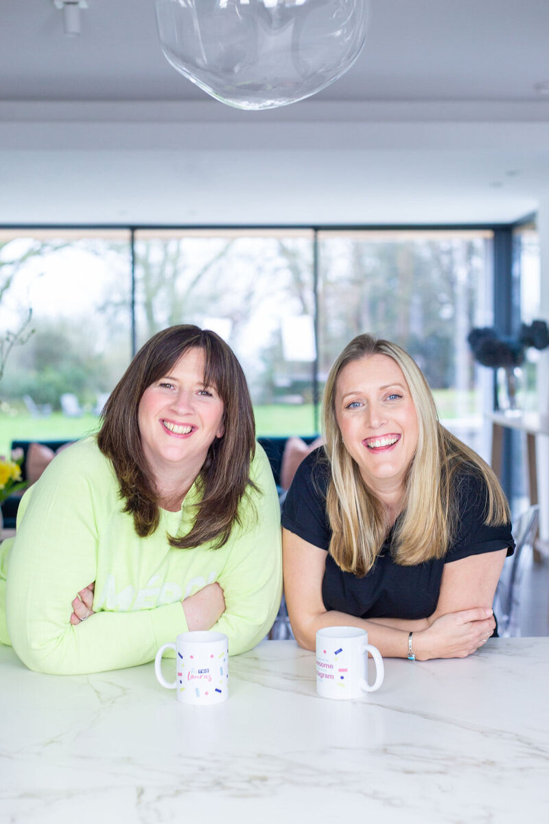 the two lauras leaning on a worktop smiling. They both have a mug in front  of them with the two lauras logo on