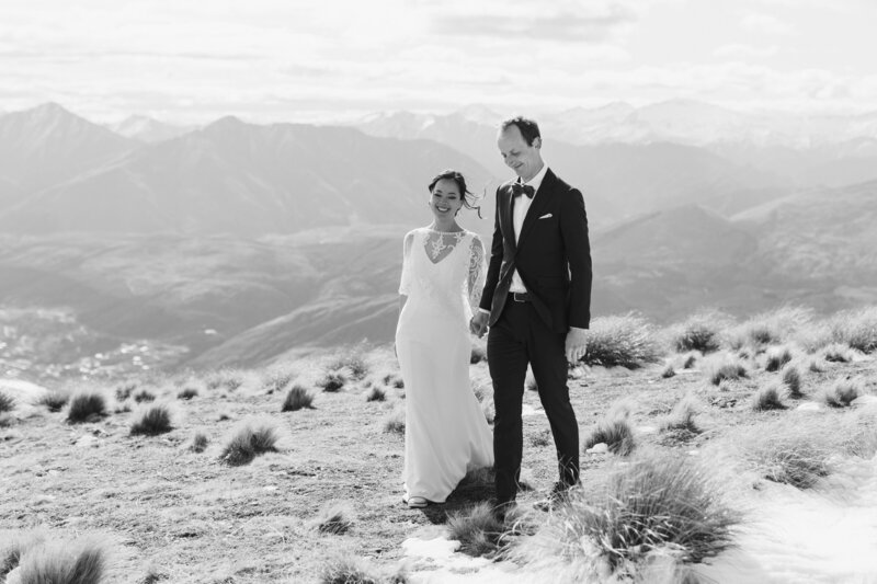 Bride and groom walking naturally hand in hand on a mountain top, the breeze is gently blowing the brides hair and they're smiling