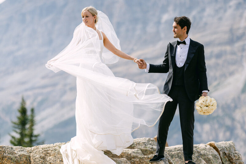 Bride and groom holding hands on rocks at Peyto Lake in Banff National Park with the bride’s veil flowing in the wind as she looks away captured as a glamorous and scenic wedding portrait by Calgary wedding photographer Geoff Wilkings