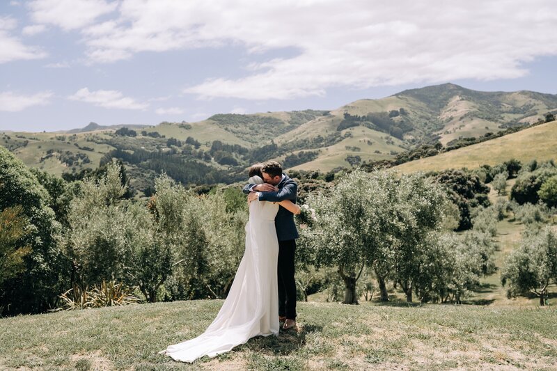 a wedding couple hug in an olive grove in akaroa new zealand overlooking mountains and valleys