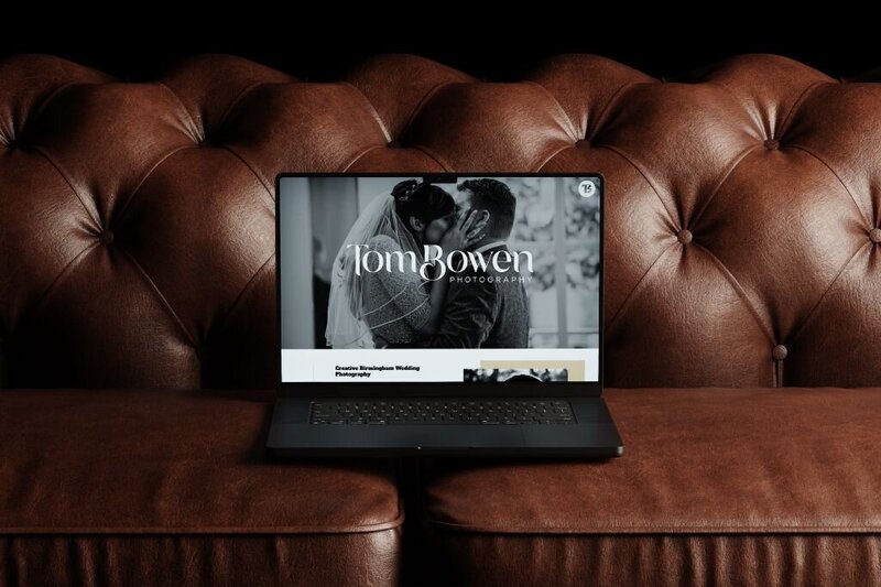 Laptop on a brown leather tufted sofa displaying a wedding photography website with a black-and-white image of a bride and groom kissing and the text “Tom Bowen Photography” prominently featured.