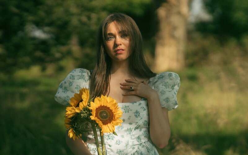 A cinematic portrayal of a young woman holding a sunflower, exuding elegance and introspection in a mystical grassland setting