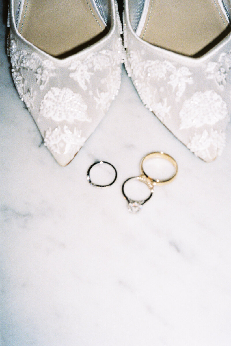 Bridal shoes and rings styled on marble– fine art flat lay.