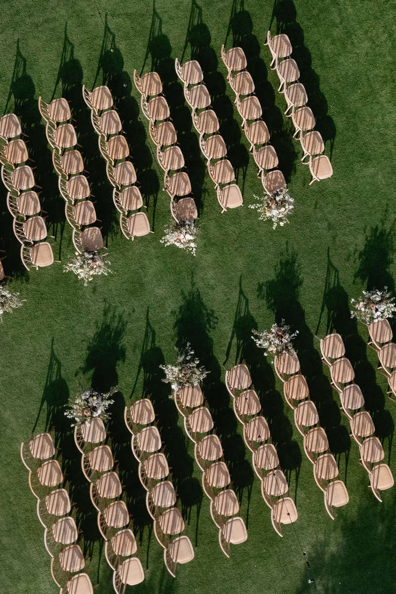 An aerial view of rows of wooden chairs arranged for an outdoor Napa Valley wedding ceremony with floral arrangements on a lush green lawn.