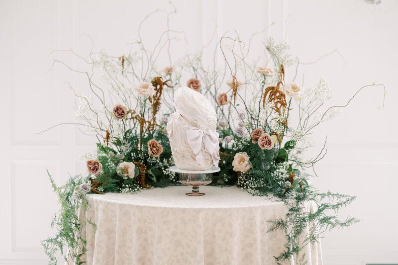 Mini foldable round table covered in a blush floral linen, styled with a sculptural wedding cake and surrounded by untamed garden-inspired florals.