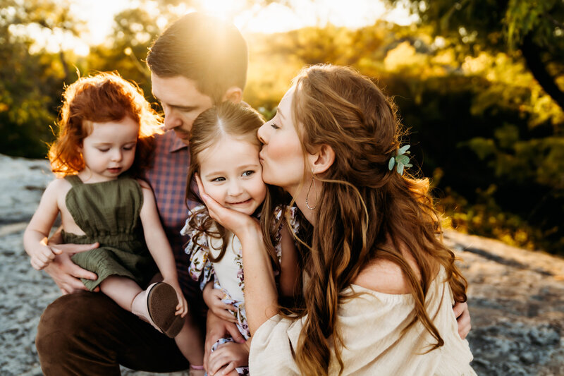 Mom kisses one daughter, other daughter sits on dad's lap