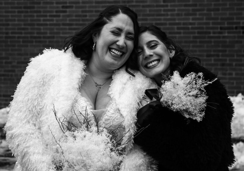 Bride and her maid of honor wearing short fur coats laughing and hugging outside in the snow