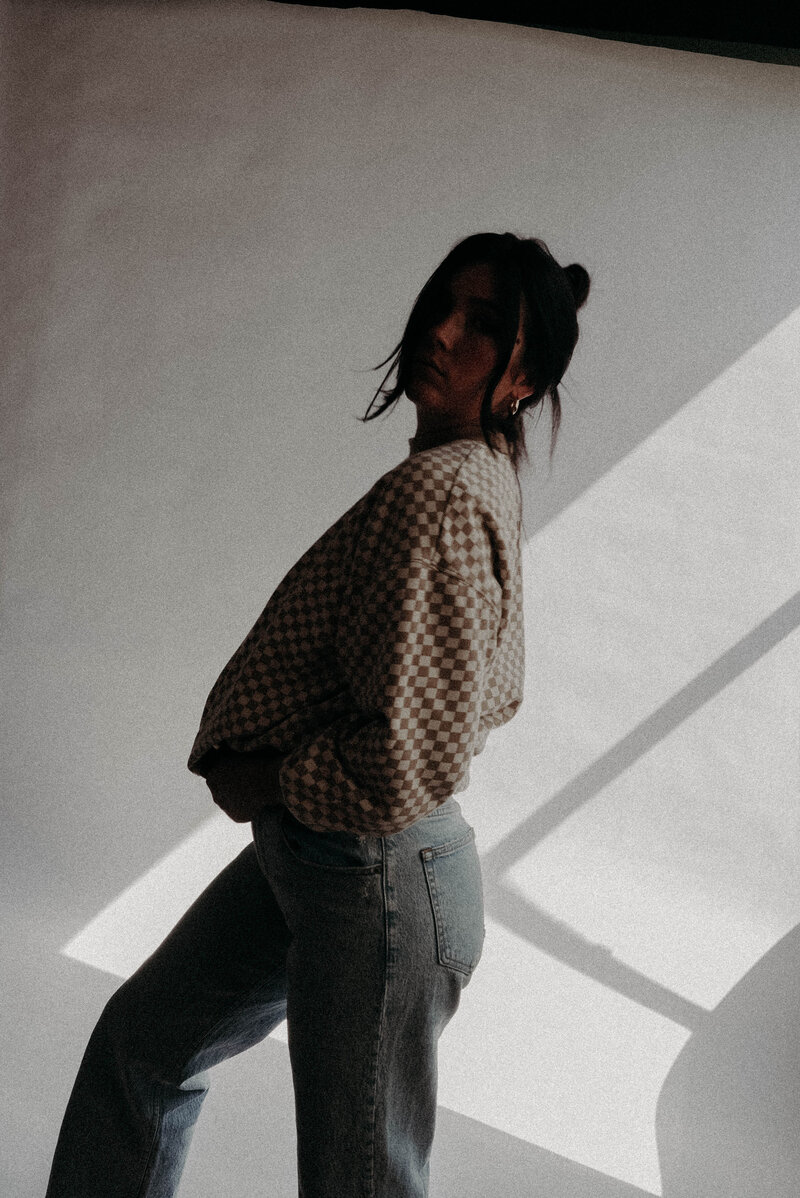 Tia Curran wearing a checkered brown sweatshirt and blue jeans sanding in front of white backdrop