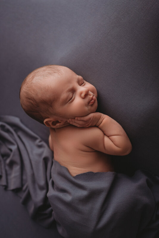 2 week newborn baby boy asleep at Atlanta, Georgia photographer's studio, laying on blue fabric backdrop, laying on tummy with gray fabric draped over his diaper and wearing a gray sleepy cap