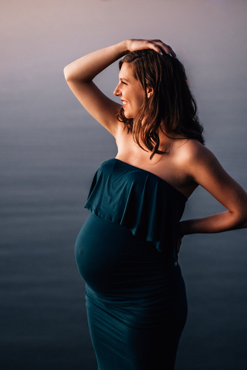 A close-up of a pregnant woman in a strapless, dark teal dress standing by a calm, foggy lake. She is smiling, with one hand resting on her hip and the other lightly touching her head.
