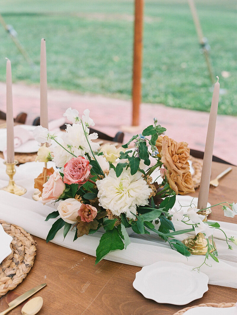 White, tan, dusty rose pink low centerpiece on long wooden table with taper candles and runner