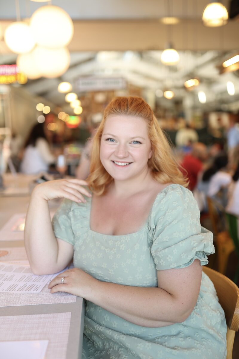 Rachel is the owner and lead content creator at Social Butterfly Wedding Content. She is photographed here in a green dress with red hair in a warm, busy space