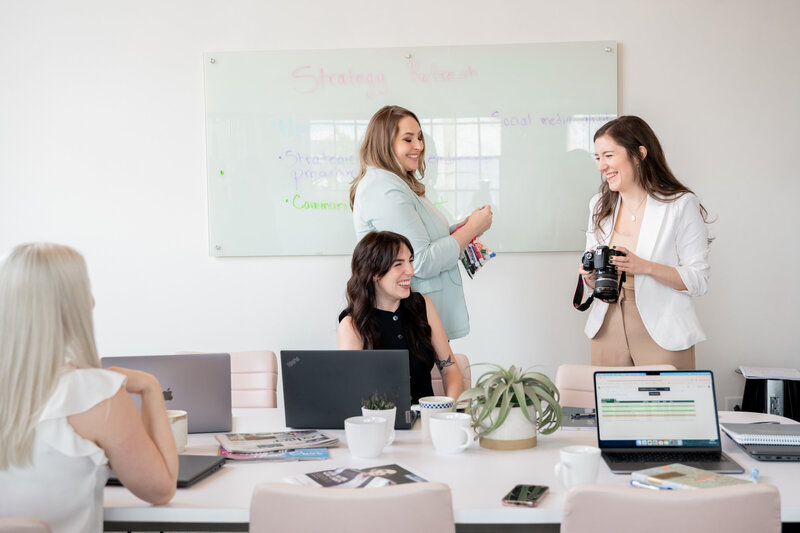 Three Pierce PR team members collaborating around a white table with laptops, notebooks, and coffee mugs, in front of large industrial-style windows.