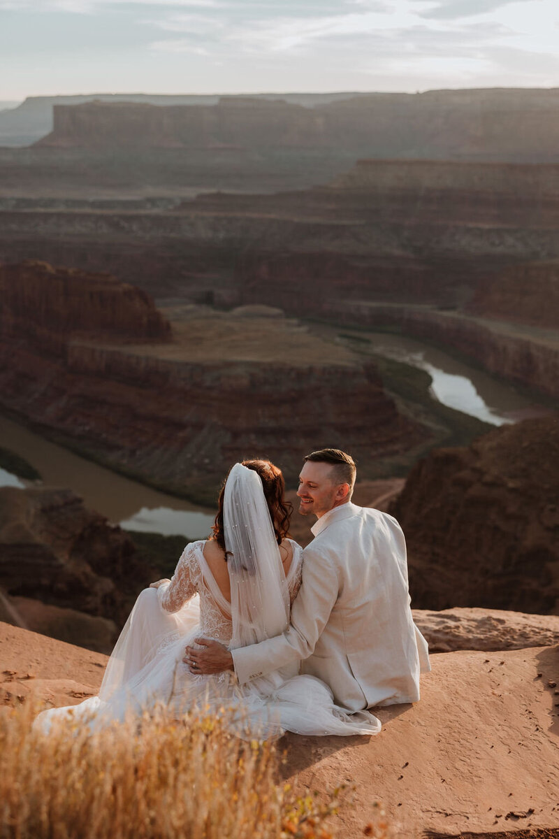 bride and groom sitting at the edge of a cliff in the desert with a winding river in the back ground