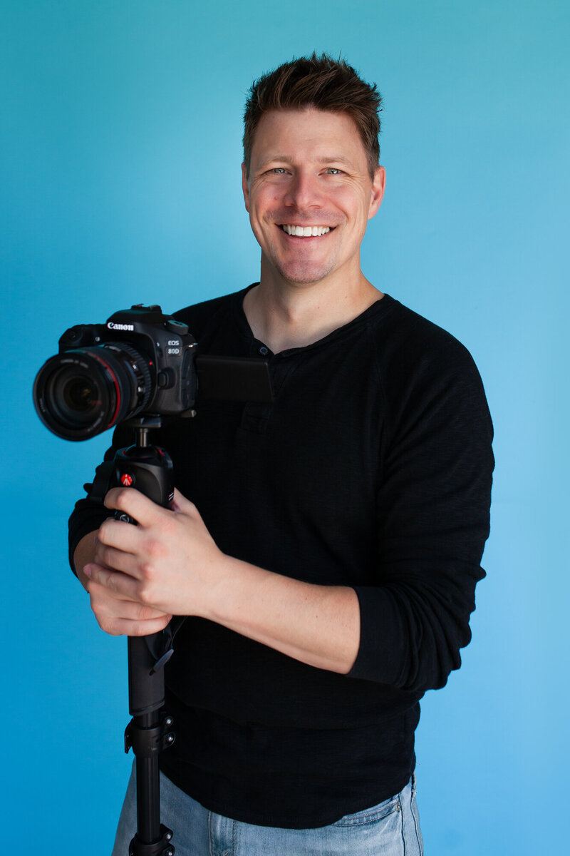 Smiling headshot of Matthew Levingston with his camera in a black shirt in front of a seamless turquoise backdrop