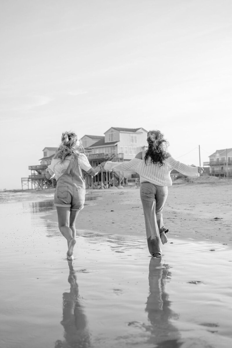 a black and white photo of two friends running in the ocean barefoot with arms outstretched