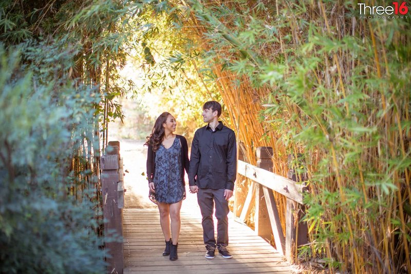 Engaged couple walk hand in hand across a bridge amongst plenty of plants at the Fullerton Arboretum