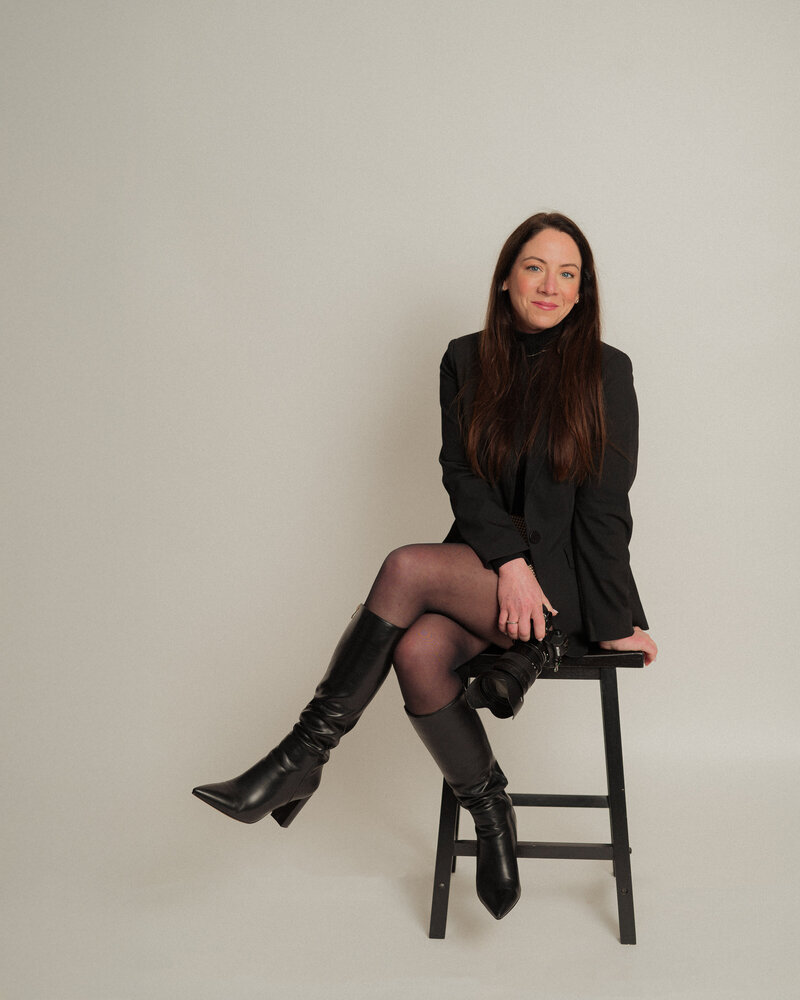 A woman with long brown hair, dressed in a black blazer, skirt, tights, and boots, sits on a tall black stool against a plain light background.