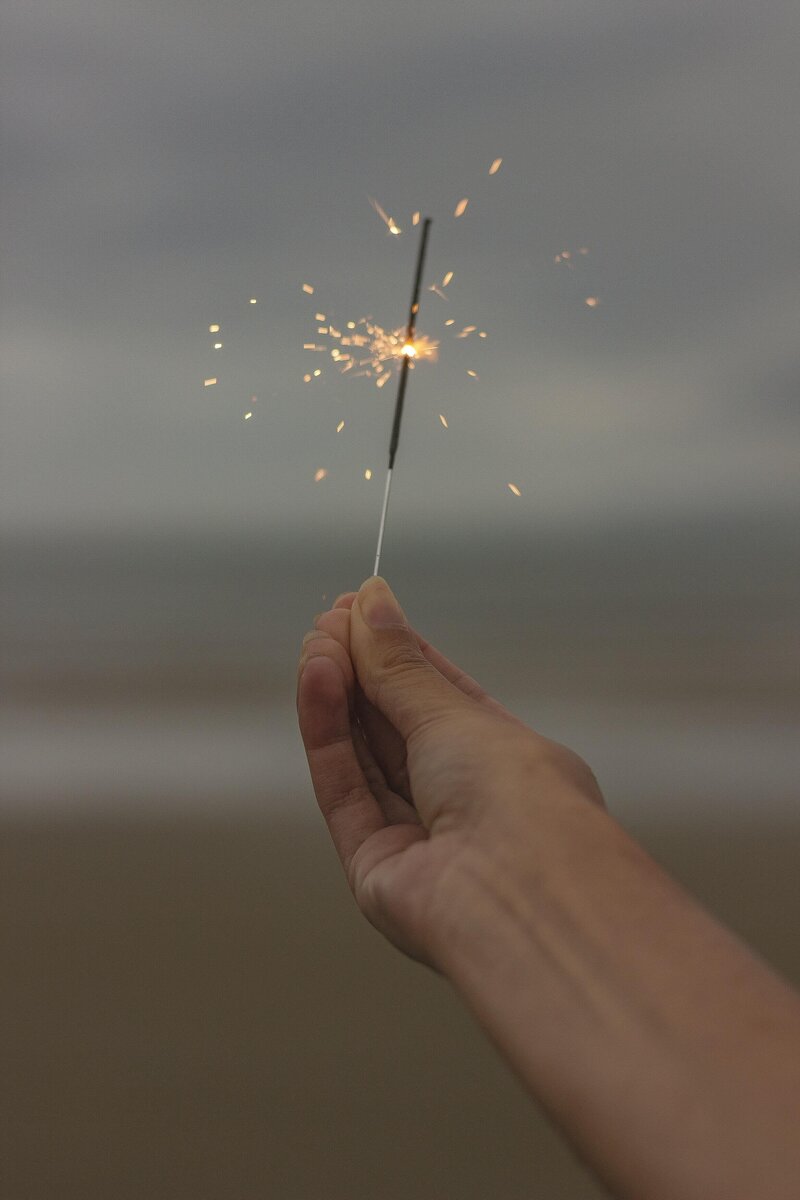 Hand, die eine funkelnde Wunderkerze hält, im Hintergrund die Umrisse des Strandes erkennbar: Innere Kraft und das Leuchten des Selbst entfachen.