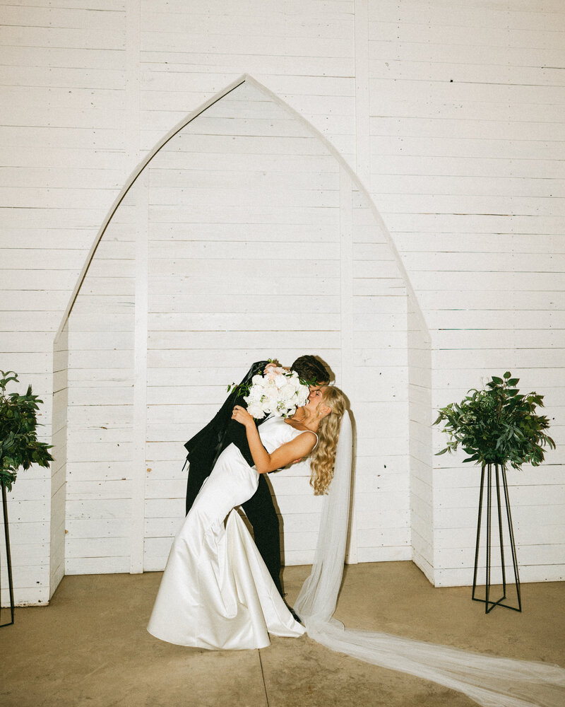 Groom in tan vest, blue tie, and pants sitting and hugging Bride