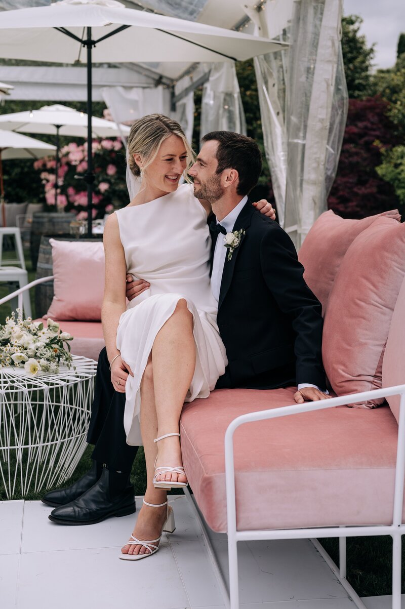 a bride sits on groom's knee on pink velvet couch from little hire company in their clear marquee with white umbrellas