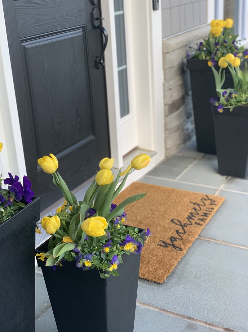 Purple flowers and yellow tulips in planter in front of door mat and black front door