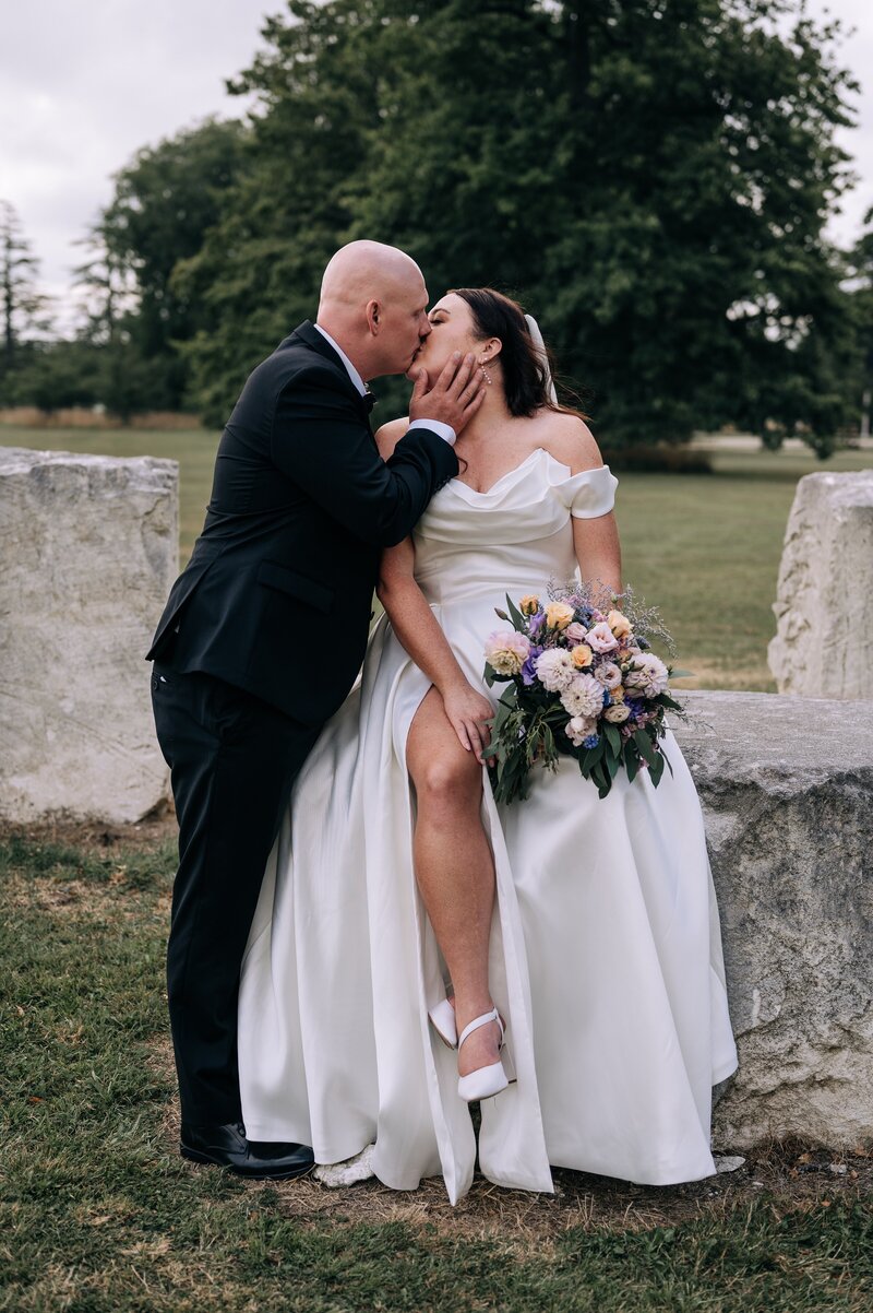 bride in paperswan dress sits on stone block at bangor farm while groom kisses her