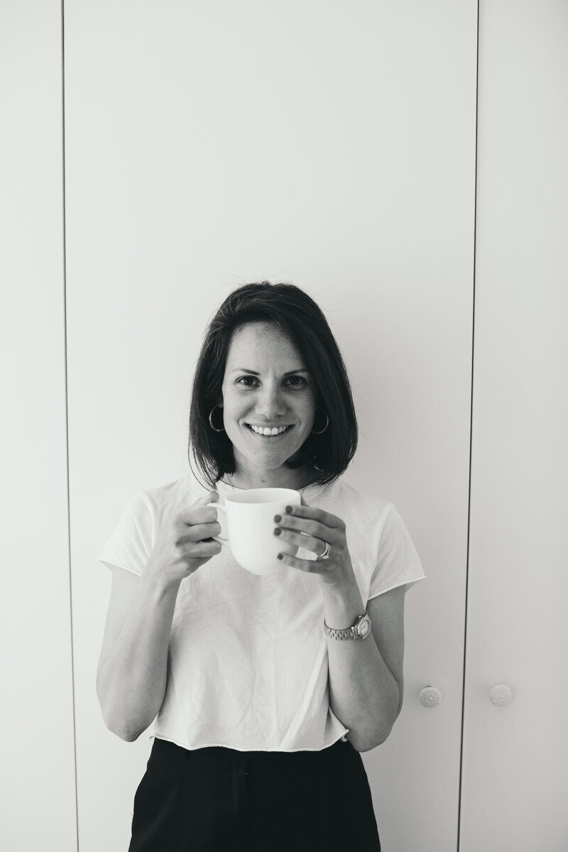 Black-and-white photo of a smiling Kaya Ramjee in a white t-shirt, holding a mug with both hands