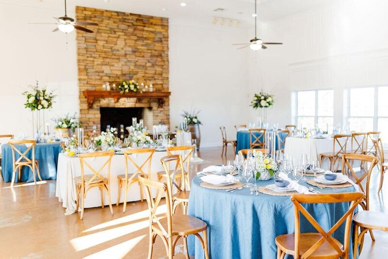 A beautifully arranged wedding reception with blue linens, cross-back chairs, and floral centerpieces, set against a stunning stone fireplace at Willow and Stone Events near Charlotte, NC.